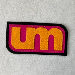 May include: A black felt patch with a pink background and the letters "um" in yellow felt.