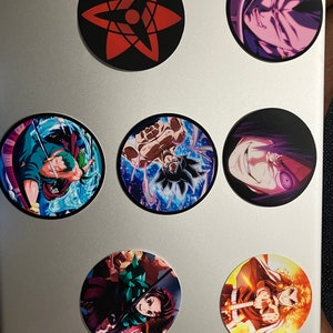 May include: A collection of round stickers featuring anime characters and symbols. The stickers showcase vibrant colors and detailed illustrations of various characters and designs, including a red and black starburst symbol.