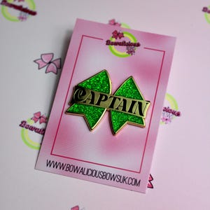 May include: A green glittery bow-shaped pin with gold trim and a central gold bar featuring the word CAPTAIN in black lettering. The pin is mounted on a rectangular pink display card with the text Bowalicious Bows and the website URL.