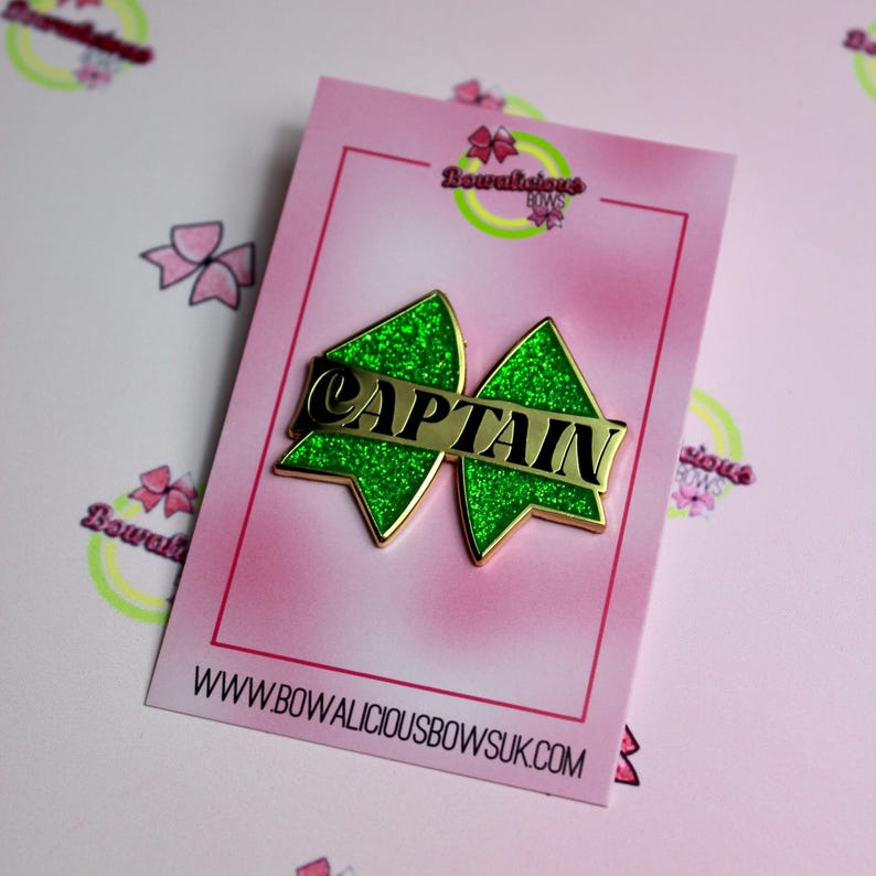 May include: A green glittery bow-shaped pin with gold trim and a central gold bar featuring the word CAPTAIN in black lettering. The pin is mounted on a rectangular pink display card with the text Bowalicious Bows and the website URL.