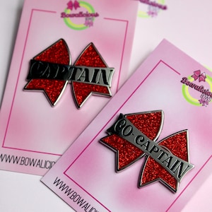 May include: Two red glitter bow-shaped pins on pink display cards. One pin features the silver text CAPTAIN, and the other features the silver text CO-CAPTAIN. Each card displays the Bowalicious Bows logo and the website URL at the bottom.