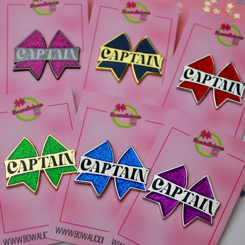 May include: Six bow-shaped pins with glittery surfaces in pink, dark blue, red, green, blue, and purple. Each pin features a horizontal banner across the center with the word CAPTAIN printed in black or white capital letters on a light background.
