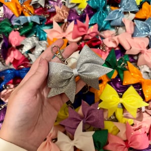 May include: A silver glitter bow keychain with a silver metal ring and clasp. The bow is surrounded by a variety of colourful glitter bows.