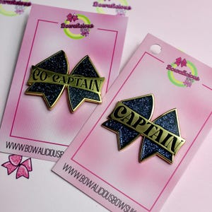 May include: Two bow-shaped enamel pins with dark blue glitter centers and gold-toned metal edges. One pin features the text CO-CAPTAIN and the other features the text CAPTAIN in black lettering on a gold banner across the center of each bow.