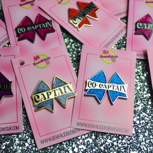 May include: A collection of glittery bow-shaped enamel pins attached to pink display cards. Each pin features the text CAPTAIN or CO-CAPTAIN in black lettering. The pins come in various colors including blue, dark blue, red, and purple with gold trim.
