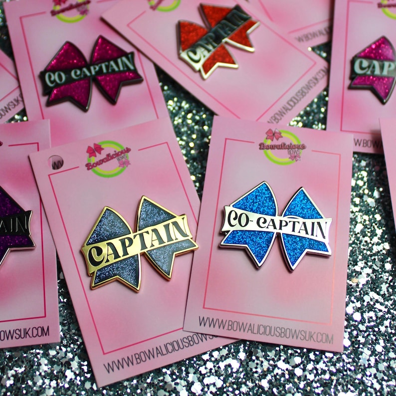 May include: A collection of glittery bow-shaped enamel pins attached to pink display cards. Each pin features the text CAPTAIN or CO-CAPTAIN in black lettering. The pins come in various colors including blue, dark blue, red, and purple with gold trim.