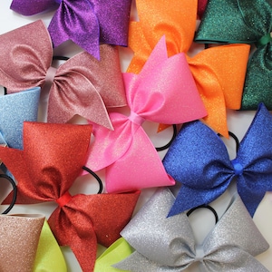 May include: A selection of glitter hair bows in shades of pink, red, purple, orange, blue, and silver. Each bow is fixed to a black elastic band. The bows are arranged closely together, displaying their vibrant colours and textures.