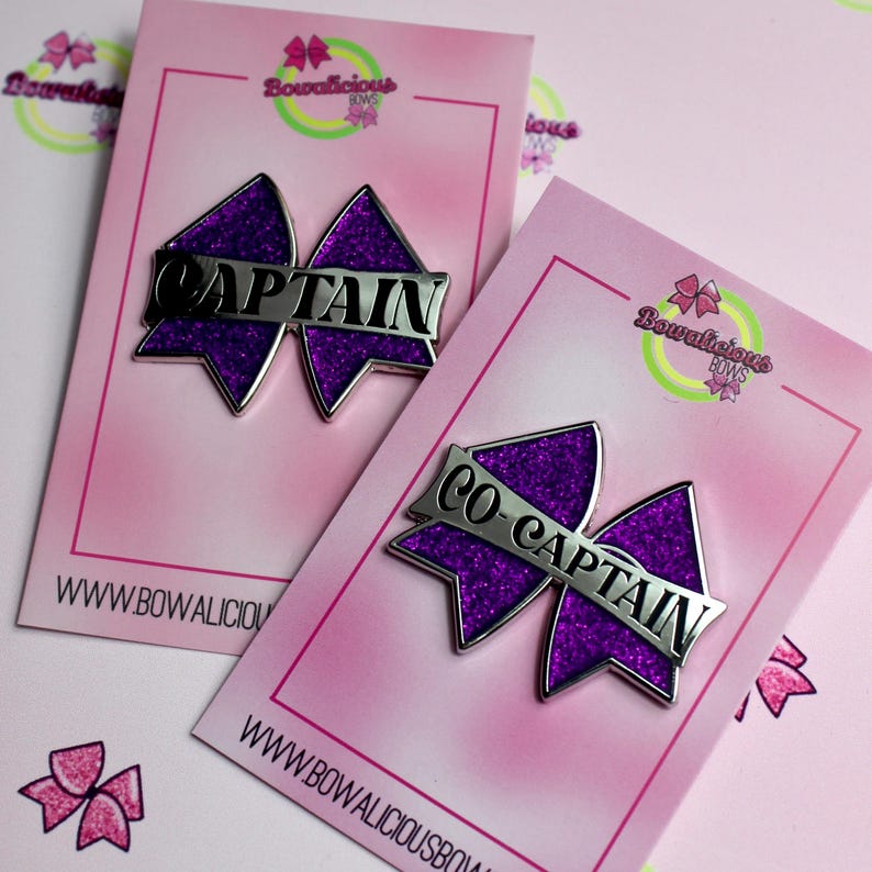 May include: Two purple glitter bow-shaped pins on pink display cards. One pin features the silver text CAPTAIN, and the other features the silver text CO-CAPTAIN. Each card displays the Bowalicious Bows logo and website address at the bottom.
