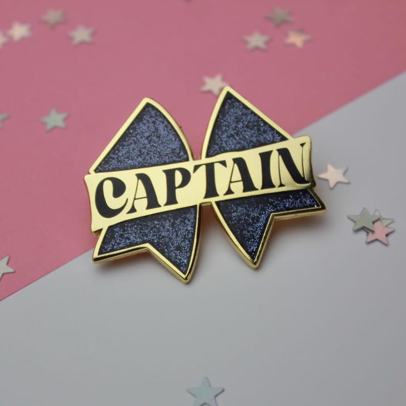 May include: A bow-shaped enamel pin with a dark blue glitter finish and gold-toned metal edges. A horizontal gold banner across the center features the word CAPTAIN in bold black capital letters. The design mimics a cheerleading hair bow.