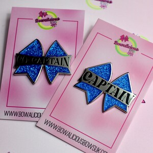 May include: Two blue glitter bow-shaped enamel pins attached to pink display cards. One pin features the silver text CO-CAPTAIN, and the other features the silver text CAPTAIN. The cards display the Bowalicious Bows logo and website address.