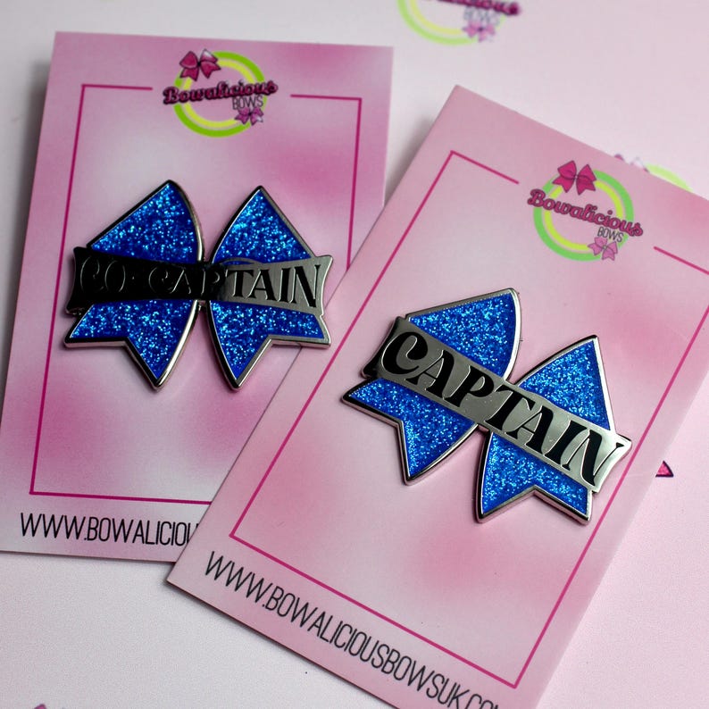 May include: Two blue glitter bow-shaped enamel pins attached to pink display cards. One pin features the silver text CO-CAPTAIN, and the other features the silver text CAPTAIN. The cards display the Bowalicious Bows logo and website address.