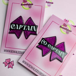 May include: Two bow-shaped pins on pink display cards. One pin features the word CAPTAIN in white letters on a black bar over a purple glitter bow. The other pin features the word CO-CAPTAIN in white letters on a black bar over a purple glitter bow.