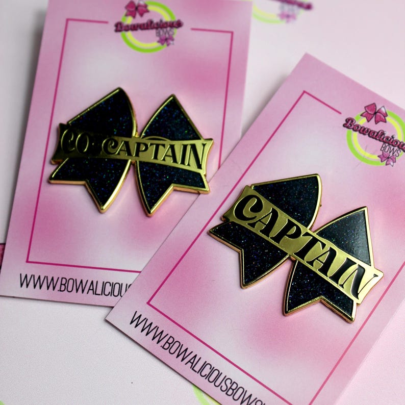 May include: Two bow-shaped pins with dark blue glitter surfaces and gold-toned metal edges. One pin features the text CO-CAPTAIN and the other features the text CAPTAIN in gold lettering. Each pin is mounted on a pink display card with Bowalicious Bows branding.
