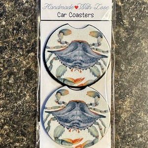May include: Two round car coasters with a blue crab design. The coasters are white with a blue crab in the center. The crab has orange claws and a white background. The text "Handmade with Love Car Coasters" is printed on the packaging.