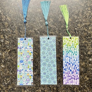 May include: Three decorative bookmarks with colorful tassel accents. The bookmarks feature winter-themed patterns, including snowflakes and illustrated forest scenes. Each bookmark is protected in a clear plastic sleeve.
