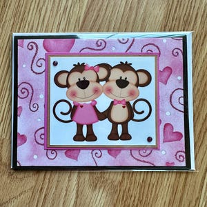 May include: A pink and white Valentine's Day card with a couple of monkeys holding hands. The monkeys are brown with pink accents and are wearing pink and brown bows. The card has a pink background with white swirls and hearts.