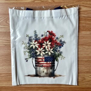 May include: A white canvas tote bag with a red, white, and blue American flag design on a metal bucket filled with red, white, and blue flowers.