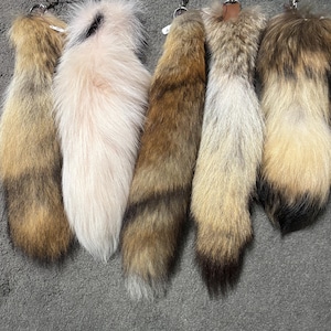 May include: Five fluffy animal tails with metal keychains attached at the top. The tails feature various natural color patterns including shades of tan, cream, brown, grey, and black, with long, soft fur textures displayed in a vertical row.