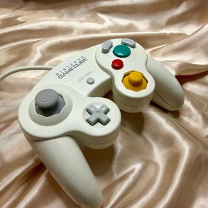 May include: A white Nintendo GameCube video game controller featuring a gray analog stick, a yellow C-stick, a green A button, a red B button, gray X and Y buttons, a gray D-pad, and a Start/Pause button with the Nintendo GameCube logo.