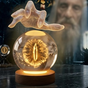 Könnte beinhalten: Eine klare Glaskugel mit einem leuchtenden, goldenen Augendesign, auf einem Holzsockel. Eine Hand schwebt über der Kugel. Der Hintergrund zeigt ein verschwommenes Bild einer bärtigen Figur.