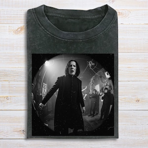 May include: Dark gray t-shirt featuring a black and white image of a man in a long black coat, standing in a dimly lit room. The image is centered on the shirt, with the neckline visible above.