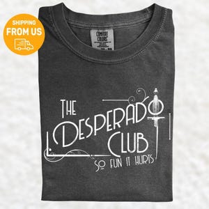Könnte beinhalten: Dunkelgraues T-Shirt mit dem weißen Schriftzug "The Desperado Club" und einer Schwertillustration. Der Satz "So Fun It Hurts" ist ebenfalls auf dem T-Shirt aufgedruckt. Das T-Shirt besteht aus einem weichen Material.