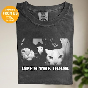 Könnte beinhalten: Dunkelgraues Comfort Colors T-Shirt mit einer Schwarz-Weiß-Grafik von Katzen. Die Grafik zeigt drei Katzen, von denen eine eine Kapuze trägt, und den Text "OPEN THE DOOR" in Weiß.