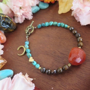 Op de afbeelding: Een turquoise en bruine kralen armband met een grote carneol hart hanger en gouden accenten. De armband heeft een halve maan hanger en kleine hart hangers.