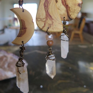 May include: Three wooden pendants with clear quartz crystals hanging from them. The pendants are made of wood with a natural grain pattern and are attached to a brown cord. The crystals are wrapped in wire.