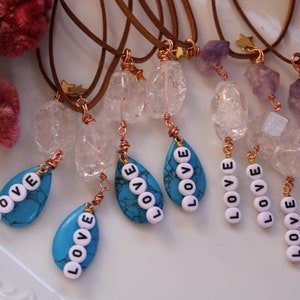 May include: A collection of eight necklaces with various crystal and gemstone pendants. Each necklace features a brown leather cord and a gold star charm. The pendants include clear quartz, amethyst, and turquoise stones. The turquoise stones have the word "LOVE" spelled out in white beads.