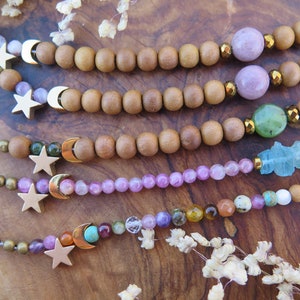 May include: Five bracelets with wooden beads, gemstone beads, and gold star and crescent moon charms. The bracelets are layered on a wooden surface.