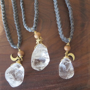 May include: Three necklaces with gray braided cords and clear quartz crystal pendants. Each pendant has a gold wire wrap and a small gold charm, either a crescent moon or a star. The cords are tied with a wooden bead.