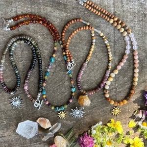 May include: An overhead shot displays several beaded necklaces with sun pendants. The necklaces feature various colors of beads, including brown, pink, and green. Some necklaces have a silver clasp. The necklaces are arranged on a wooden surface, with flowers and crystals.
