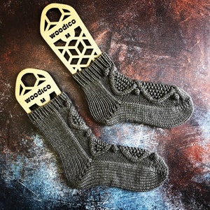 May include: Two pairs of grey knitted socks with a cable pattern and decorative bobbles, displayed on wooden sock blockers labelled "woodico M"