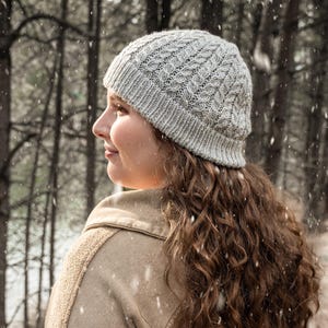 May include: A light gray knit hat with a cable knit design. The hat is worn by a person with long, curly brown hair, wearing a tan jacket. The background features a snowy forest scene.