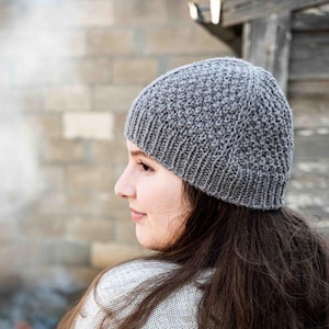 May include: A gray knitted beanie with a textured pattern. The beanie is worn by a person with long, dark brown hair and a light gray sweater. The background includes a brick structure and a fire.