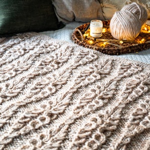 Puede incluir: Una acogedora escena de dormitorio con una manta de punto blanco, dos ovillos de lana crema, una vela y luces de cadena en una bandeja tejida. La cama está hecha con una almohada verde y una almohada gris claro.