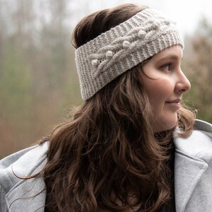 May include: A woman wearing a gray knitted headband with white pom-poms. The headband has a cable knit pattern. She is wearing a gray coat and a black shirt.