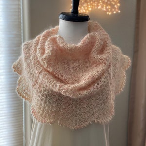 May include: A delicate, pale pink knitted shawl with a lacy pattern, displayed on a mannequin. The shawl is draped over the mannequin's shoulders and has a scalloped edge. The background features a wall with a string of lights and a pair of knitting needles crossed behind the mannequin.