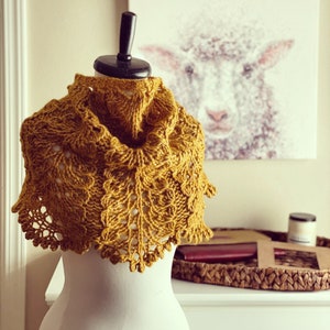 May include: A yellow knitted shawl with a delicate lace pattern, displayed on a white mannequin. The shawl is draped over the mannequin's shoulders and neck, showcasing its intricate design.