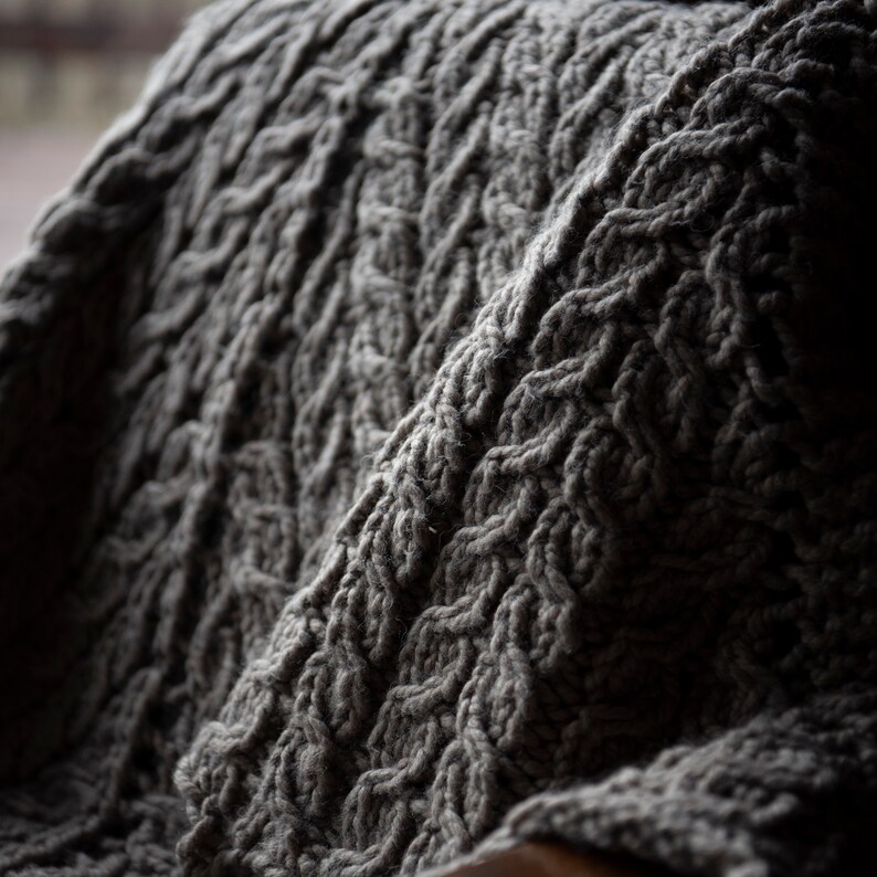 May include: A close-up of a gray knitted blanket with a cable knit pattern.
