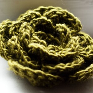 May include: A close-up shot of a handmade, olive green crocheted flower. The yarn is thick and textured, with visible stitches creating a layered, floral design. The craft item is set against a soft, white background.