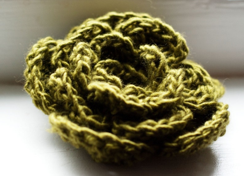 May include: A close-up shot of a handmade, olive green crocheted flower. The yarn is thick and textured, with visible stitches creating a layered, floral design. The craft item is set against a soft, white background.