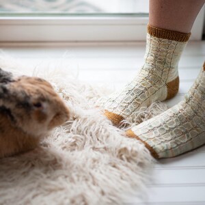 May include: A pair of white and yellow knitted socks with a textured pattern. The socks are worn by a person sitting on a fluffy white rug. A brown rabbit is sitting on the rug.