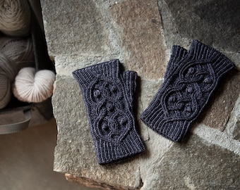 May include: A pair of dark grey fingerless gloves with a cable knit pattern and a textured design. The gloves are lying on a stone surface.