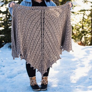 May include: A brown knitted shawl with a leaf pattern, held by a person in a snowy landscape.