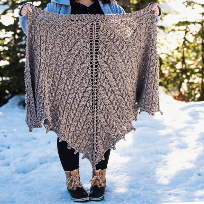 May include: A brown knitted shawl with a leaf pattern, held by a person in a snowy landscape.