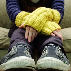 Könnte beinhalten: Eine Person trägt ein Paar gelbe, fingerlose Häkelhandschuhe und schwarze Turnschuhe. Die Handschuhe sind mit einem einfachen Stichmuster gearbeitet und haben eine leuchtend gelbe Farbe. Die Turnschuhe sind ein klassischer Stil mit einer weißen Sohle und einem schwarzen Canvas-Obermaterial.