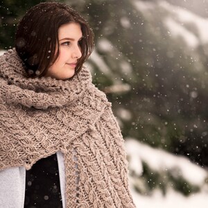 May include: A woman wearing a brown knitted scarf with a cable knit pattern. She is standing in a snowy forest with trees in the background.