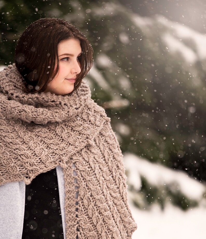 May include: A woman wearing a brown knitted scarf with a cable knit pattern. She is standing in a snowy forest with trees in the background.